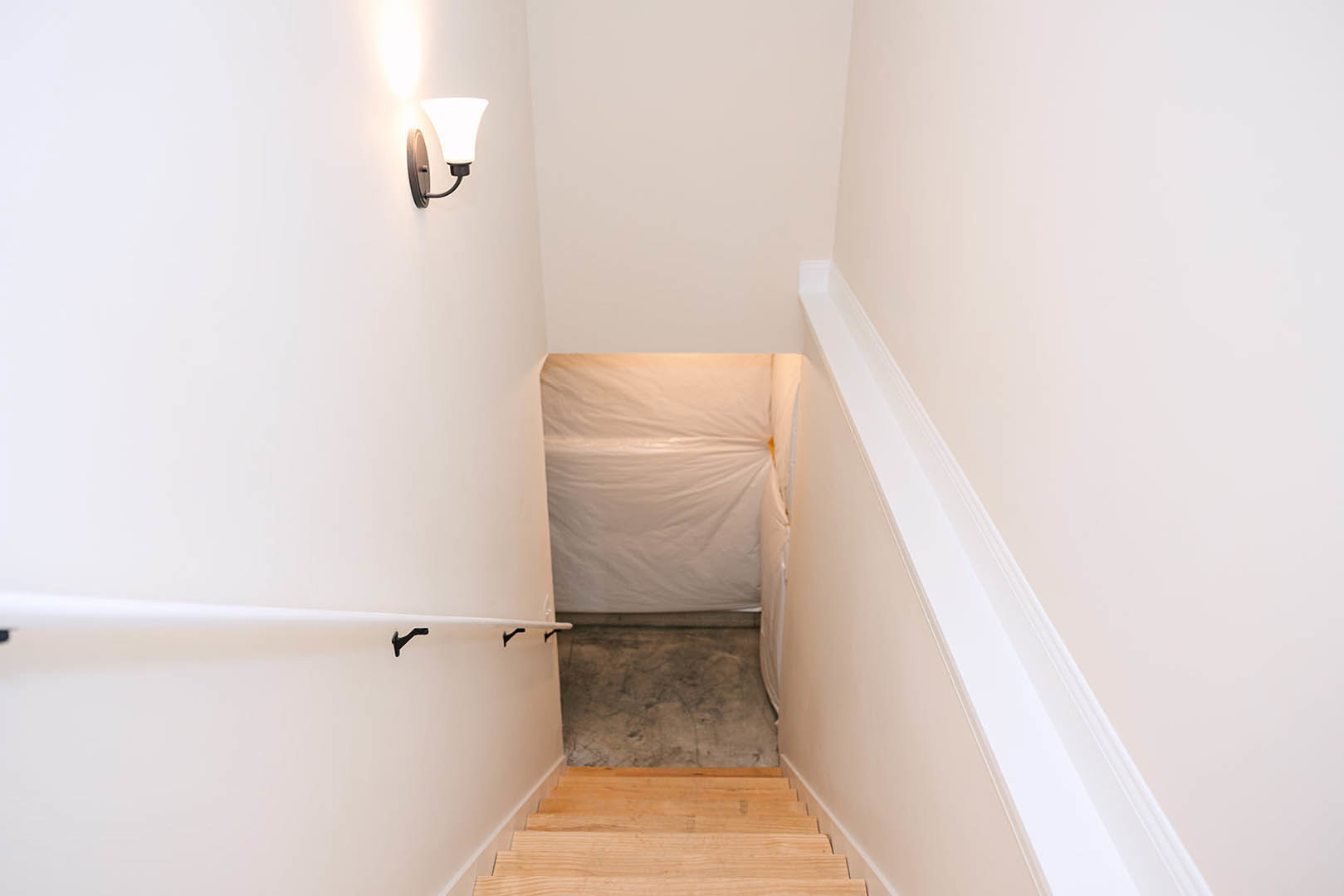 Wood staircase with white wall and sheer white curtain, light wood flooring, plaster ceiling, and modern wall lamp in a renovated indoor space.