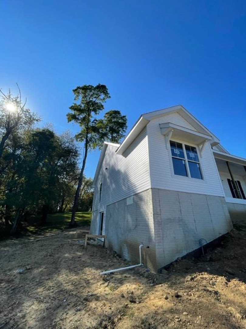 Framed custom home under construction with exposed wood, large windows, and surrounding leafy trees beneath a clear blue sky