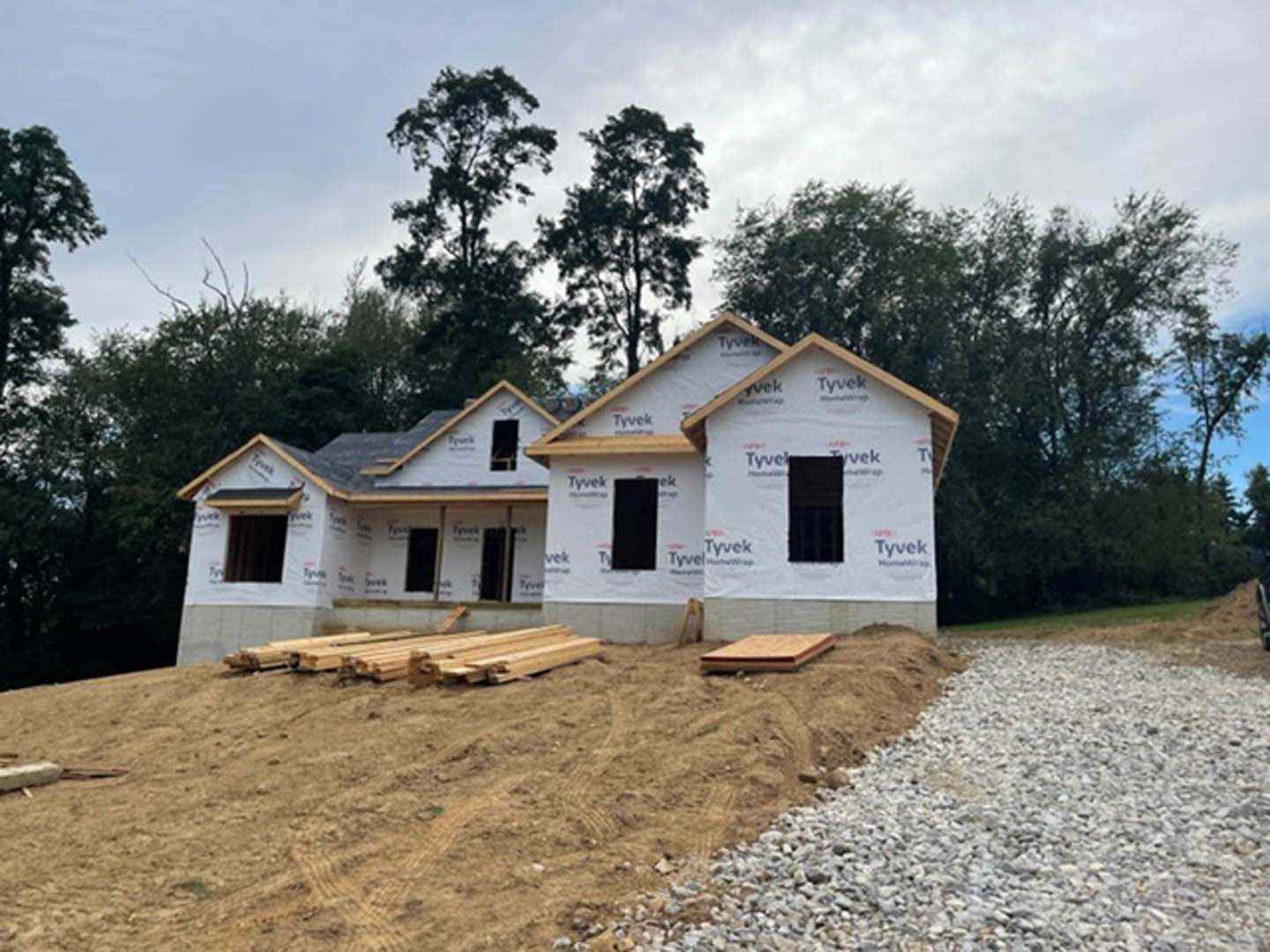 Wood-framed house under construction with exposed beams, gravel and dirt foreground, pile of lumber on ground, trees and cloudy sky in background, partially installed windows