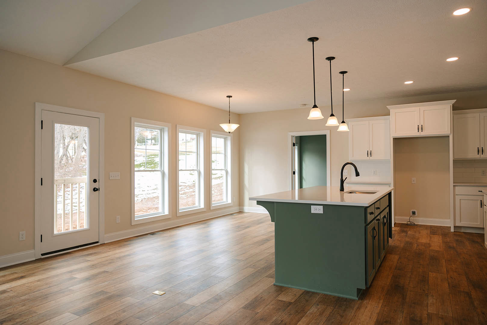 Open kitchen and living room with hardwood floors, large windows with white frames, central kitchen island featuring a sink and black pendant lamp, cabinetry along the walls