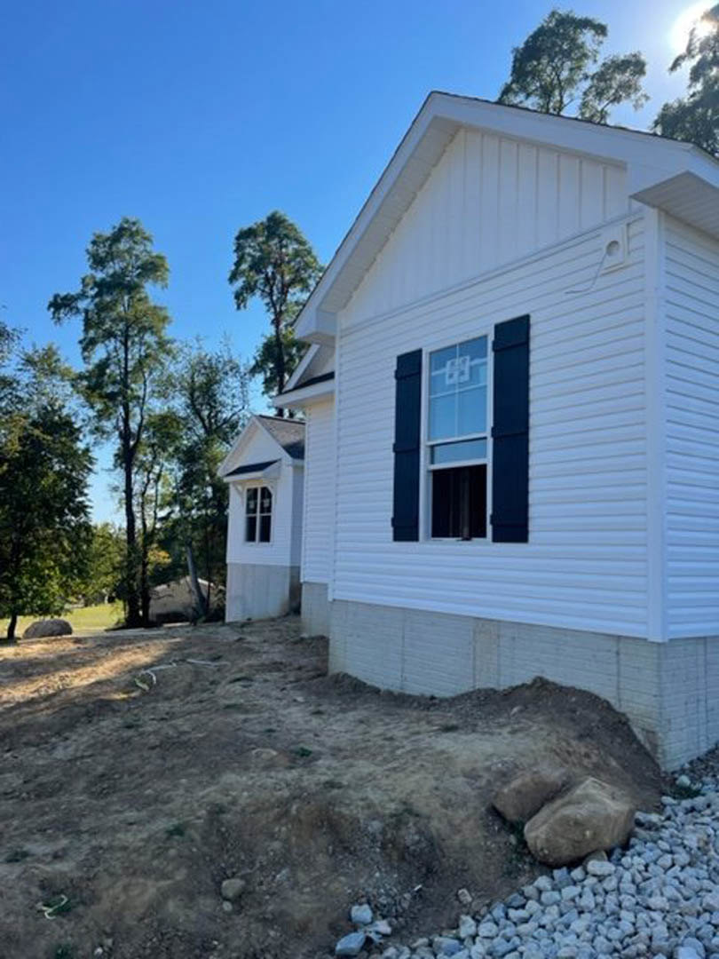 White house with black shutters, white-framed windows, blue wall with white trim, dirt and rocks on sloped ground, trees in background