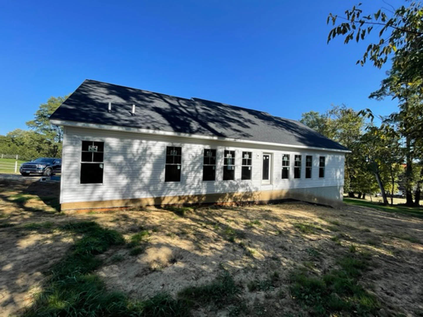 White siding house with black shingle roof, multiple rectangular windows, grassy dirt yard, trees and blue sky in background, parked car partially visible
