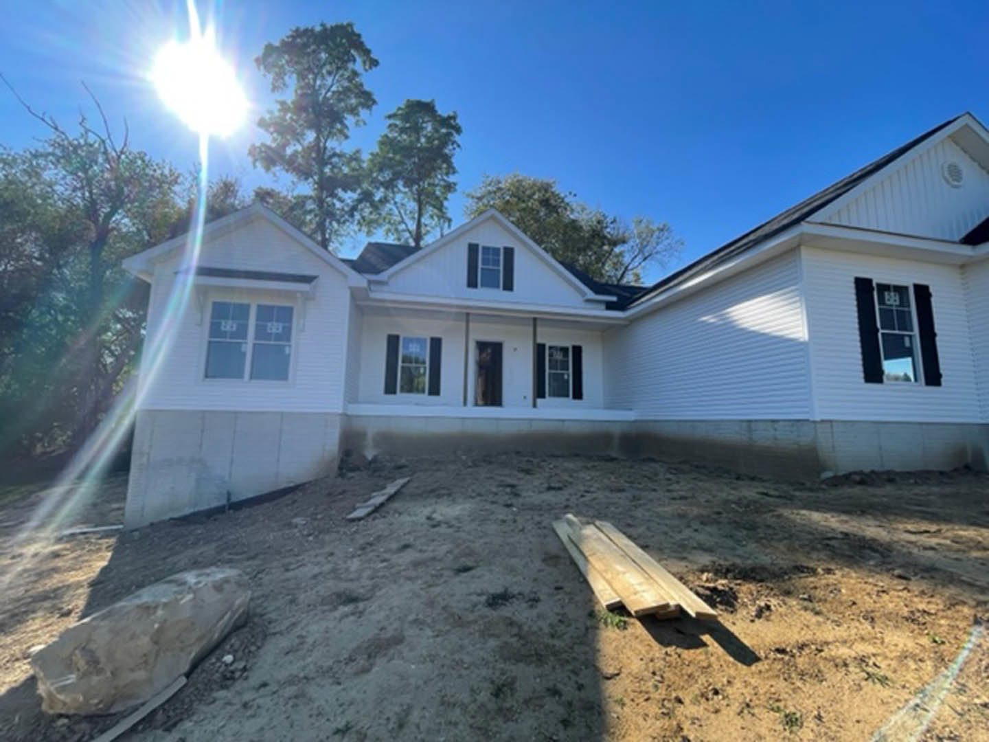 Partially built house with exposed framing, pile of lumber in front, large rock and wood plank on dirt ground, black shuttered window, trees in background