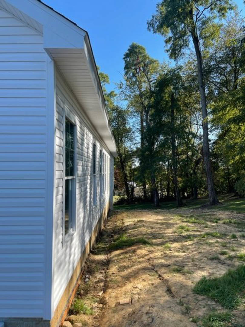 White siding house with multiple windows, surrounded by mature trees and grassy yard, dirt path leading to entrance
