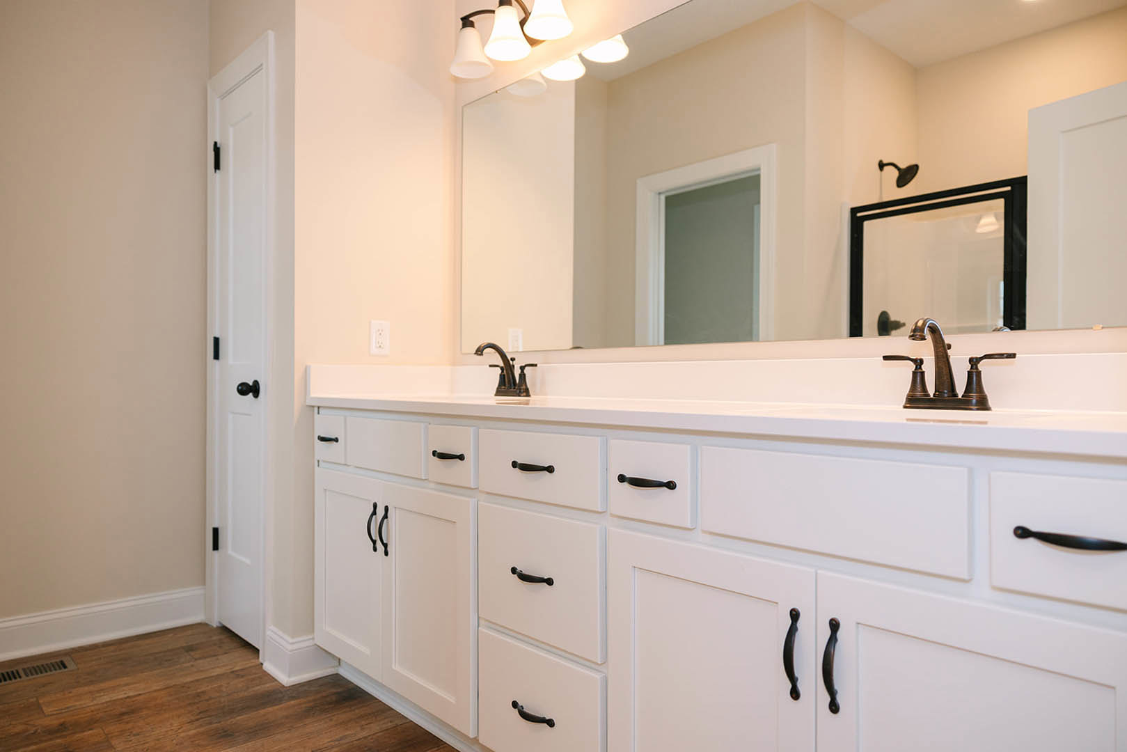 Bathroom with white shaker cabinets, quartz countertop, large framed mirror, chrome faucet, wood flooring with white baseboard trim, and tiled shower enclosure