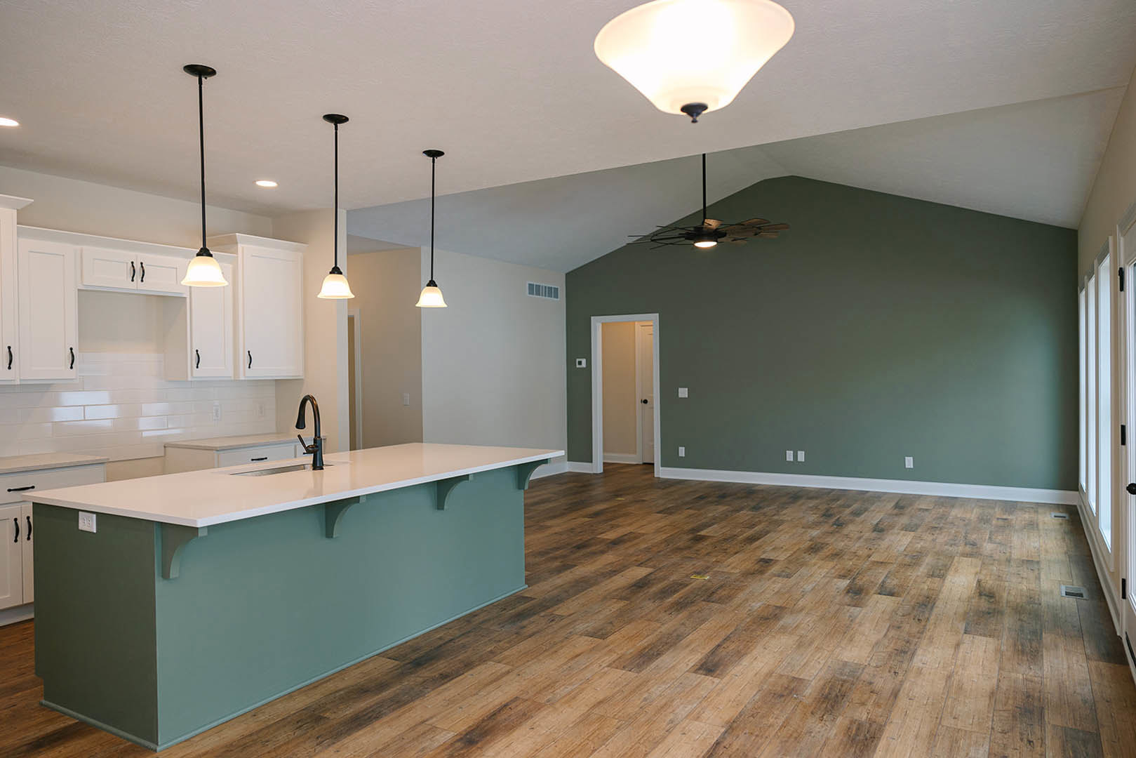 Open-concept kitchen and living room featuring wood flooring, kitchen island with sink, ceiling fan, white cabinetry, and modern light fixtures.