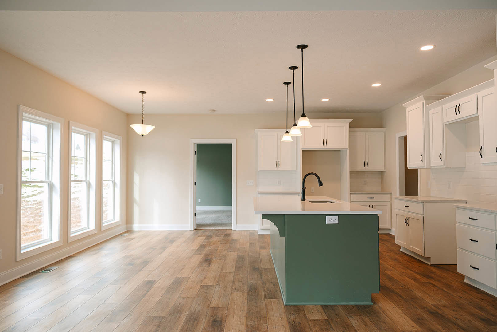 Green kitchen island with white countertop, wood flooring, white cabinetry with black handles, row of windows along wall, recessed ceiling lights