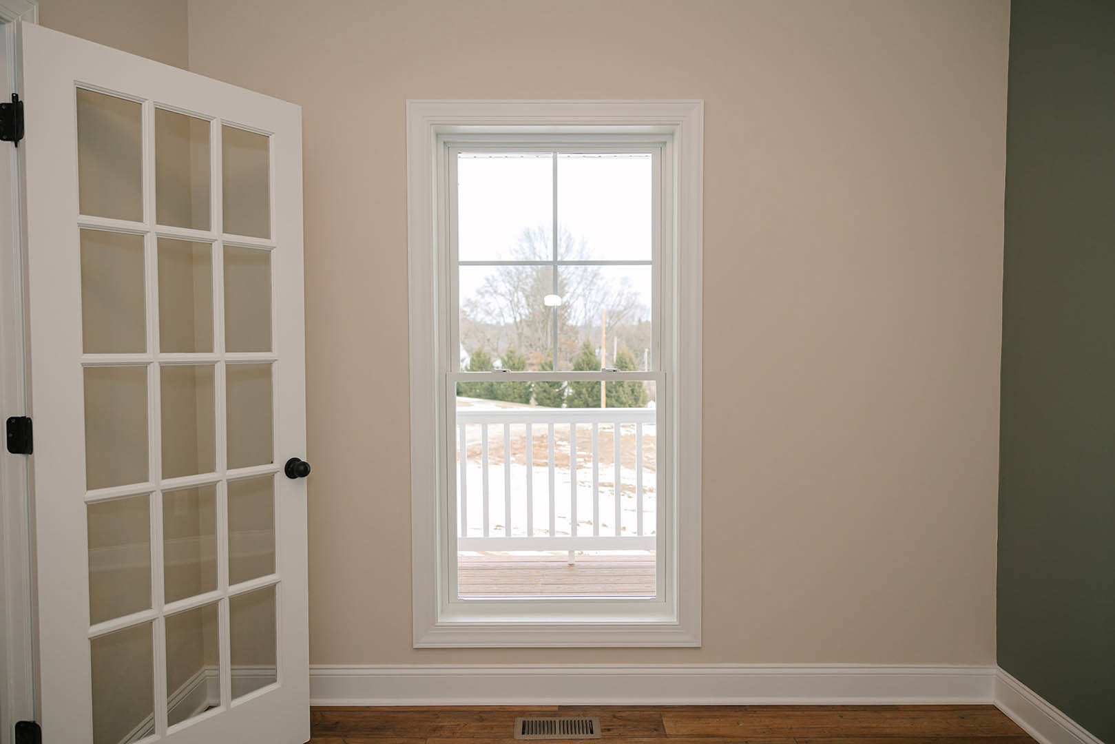 Large window with white trim overlooks snowy trees, green accent wall with white border, white door with glass panes, and white railing outside dusted with snow
