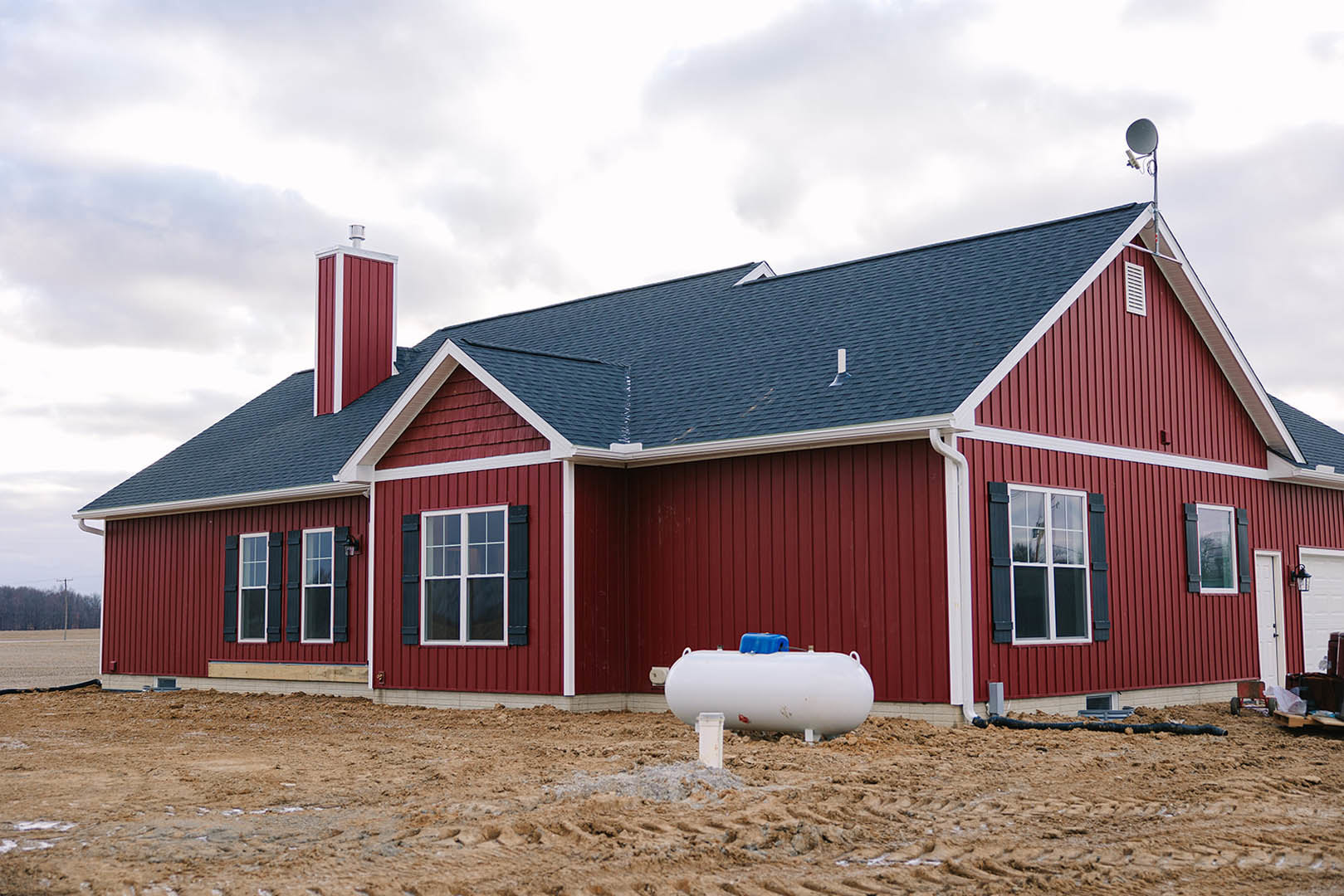 Red siding house with black shutters, white-framed windows, and a white water tank with blue top on grassy ground; pile of dirt and exposed pipe near foundation.