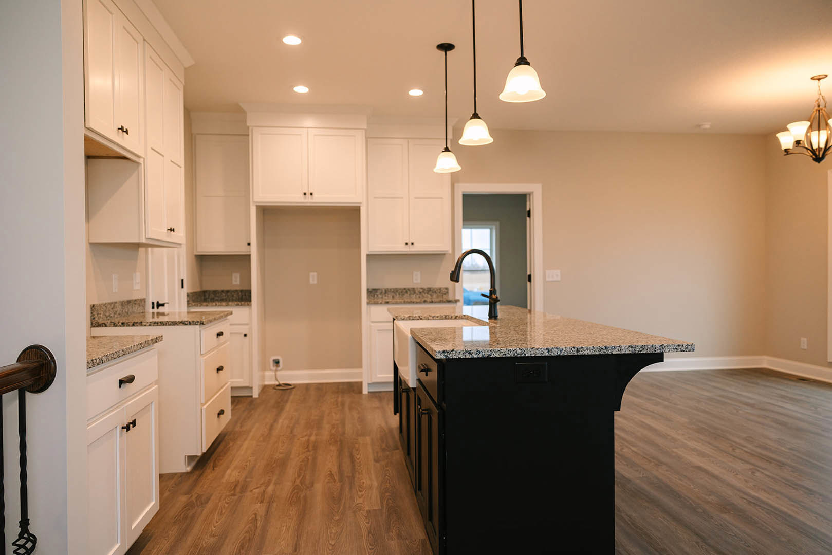 Spacious kitchen featuring a large central island with built-in sink and faucet, wood flooring, white walls with light switch, black metal pole with handle, and modern cabinetry.