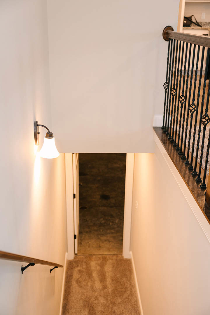 Wood staircase with black metal railing, carpeted steps, white plaster walls, hallway with paneled door, modern lamp positioned beside the stairs.