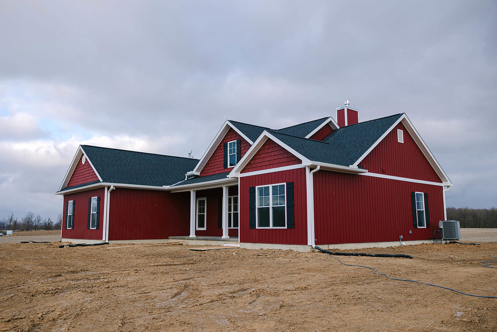 Red house with white trim under construction, close-up of window, large metal object beside red wall, dirt field in foreground, long black hose on ground, cloudy sky overhead.
