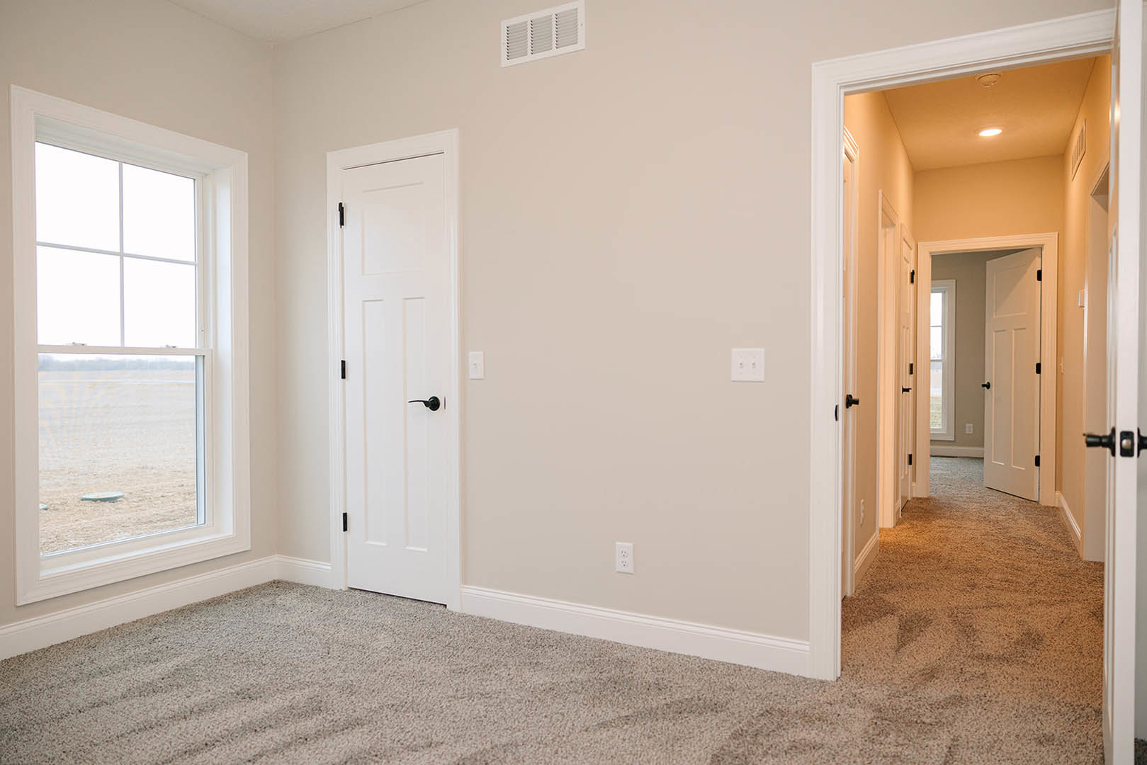 White-walled room with light carpet, large window overlooking grassy field, white door with black handle, white wall outlet with black switches, wall vent