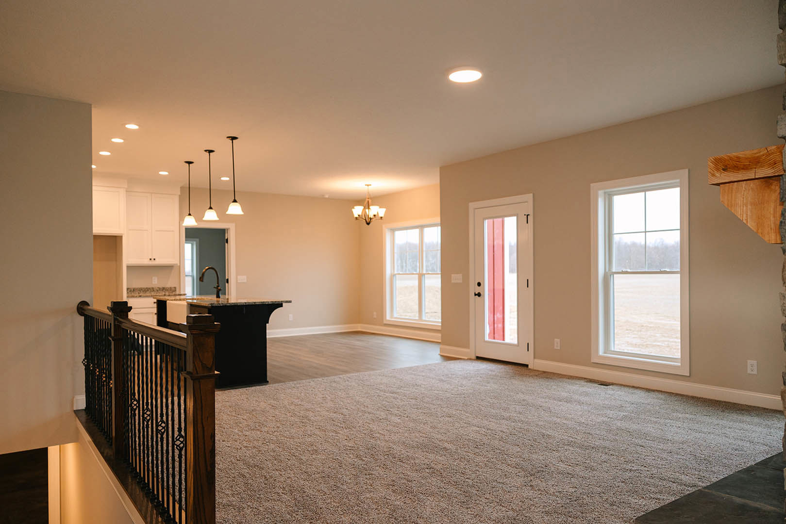 Open-concept living room with carpeted floor, adjacent kitchen featuring cabinetry and bar seating, ceiling light fixture, window overlooking a field, visible door and metal