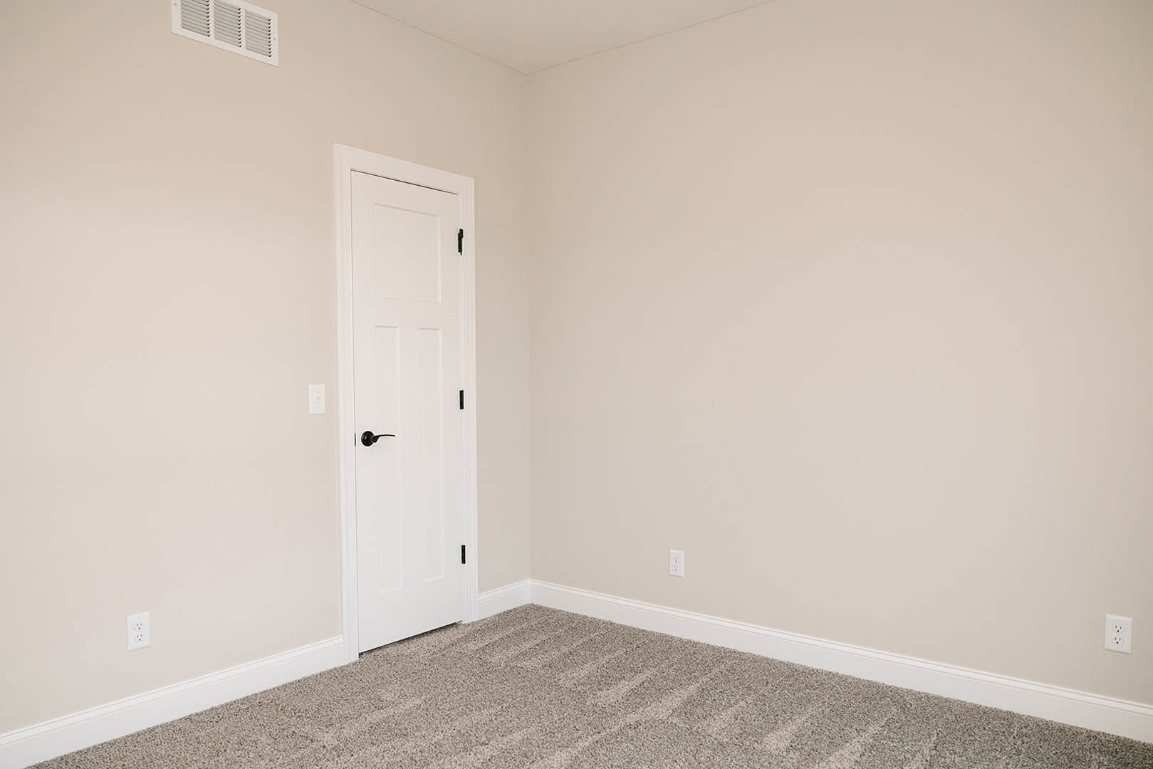 White paneled door with black handle, beige carpet flooring, white walls, ceiling vent, minimalist interior detailing