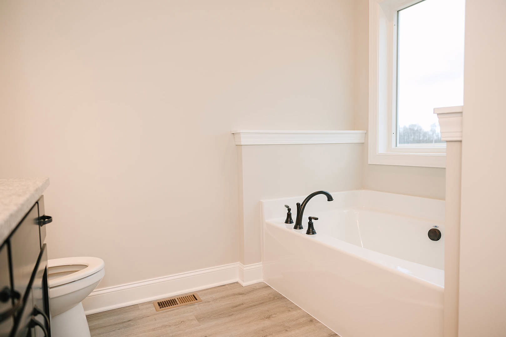 Bathroom with white tub and chrome faucets, wood flooring, close-up of white toilet, wall vent, window with white frame, and black circle with blue accent.