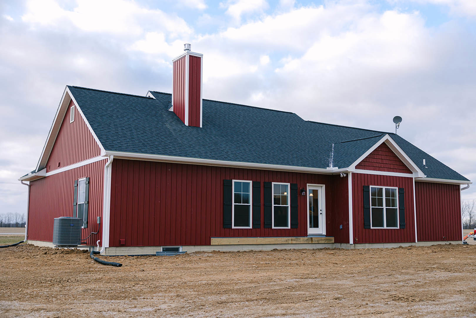 Red metal siding with white trim, white-framed window, white door with black knobs, black heat pump unit, brick chimney, cloudy sky above.