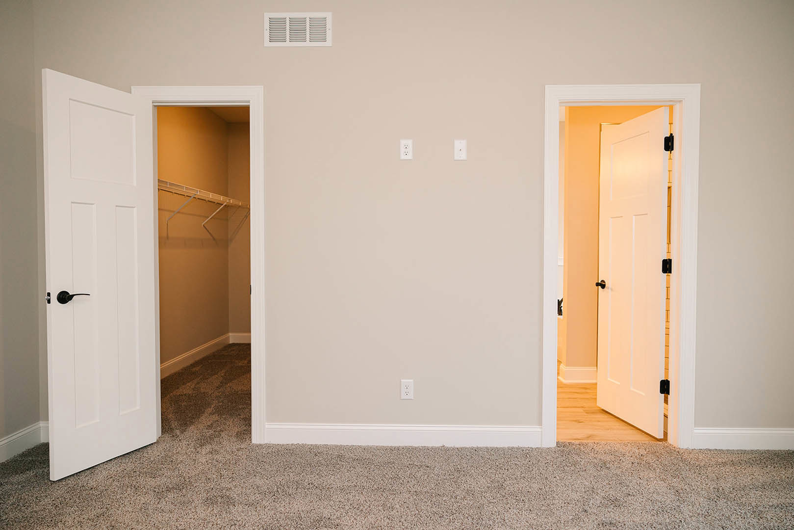White double closet doors with black handles, beige carpet flooring, white wall, and a rectangular air vent near the baseboard.