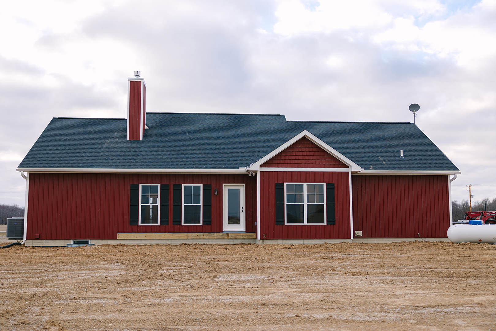 Red house with black shutters and white door featuring glass panel, set beside a dirt field under partly cloudy sky.