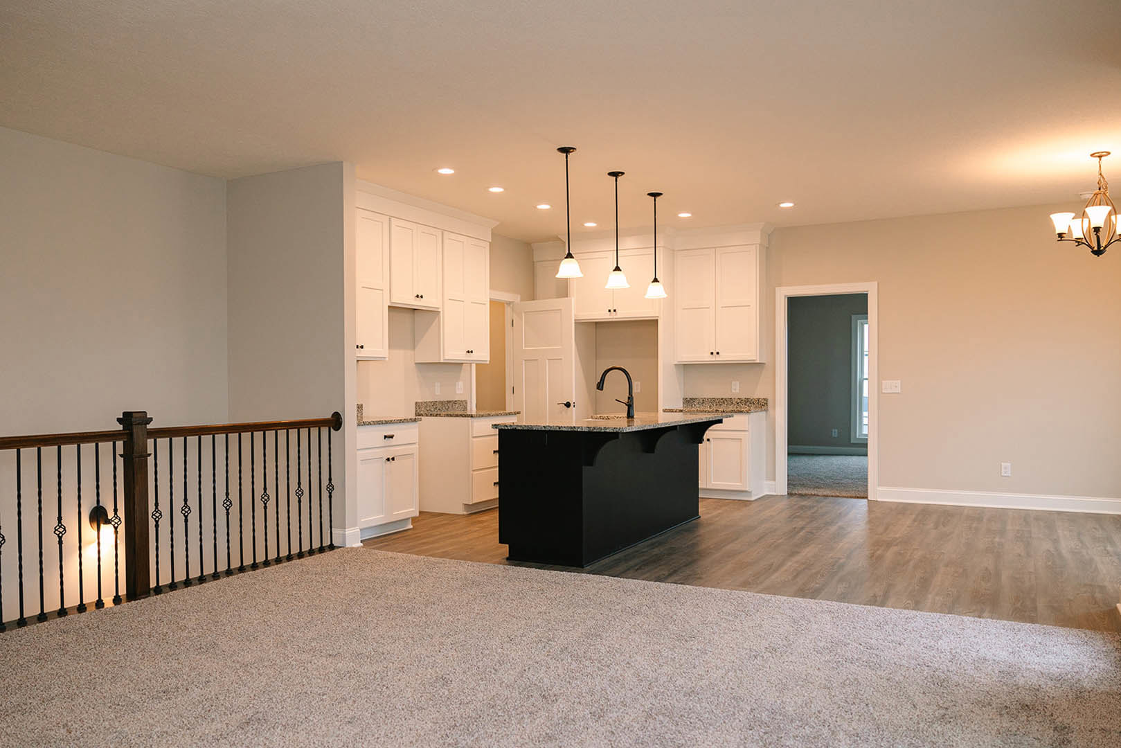 Open-concept kitchen and living room featuring a black island with marbled countertop, light wood flooring, white cabinetry, modern chandelier, and carpeted area adjacent to a