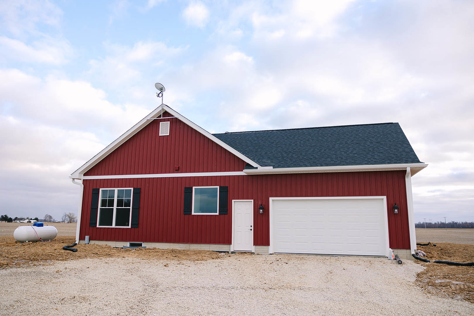 Red siding house with white garage door and red trim, white framed window, white entry door with black hardware, blue-topped white cylinder near entrance, gray shingled roof