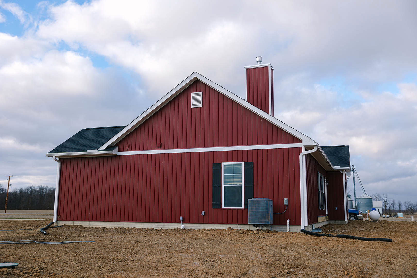 Red siding exterior with white-framed window, white vent, and brick chimney; metal utility box and round white object with blue top on dirt ground; grassy lot under partly cloudy