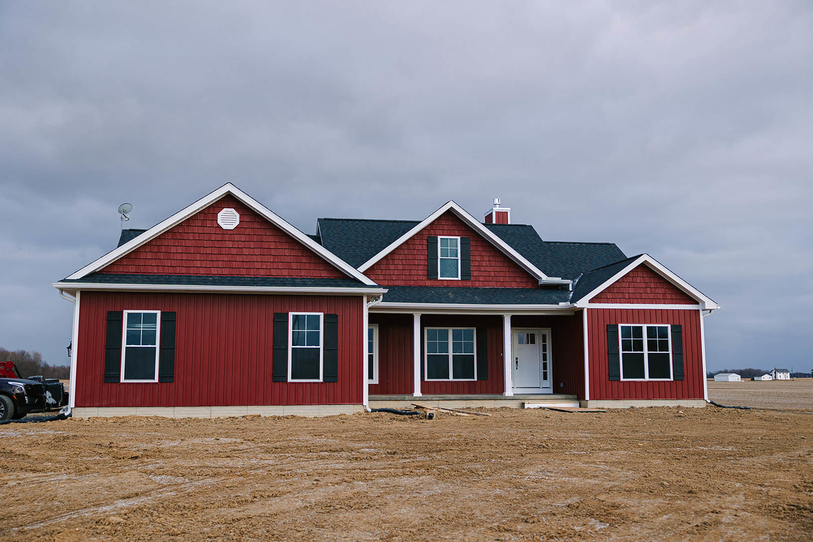 Red siding house under construction with white vent, glass-paneled white door, and white-framed window, set beside a dirt field under a cloudy sky.