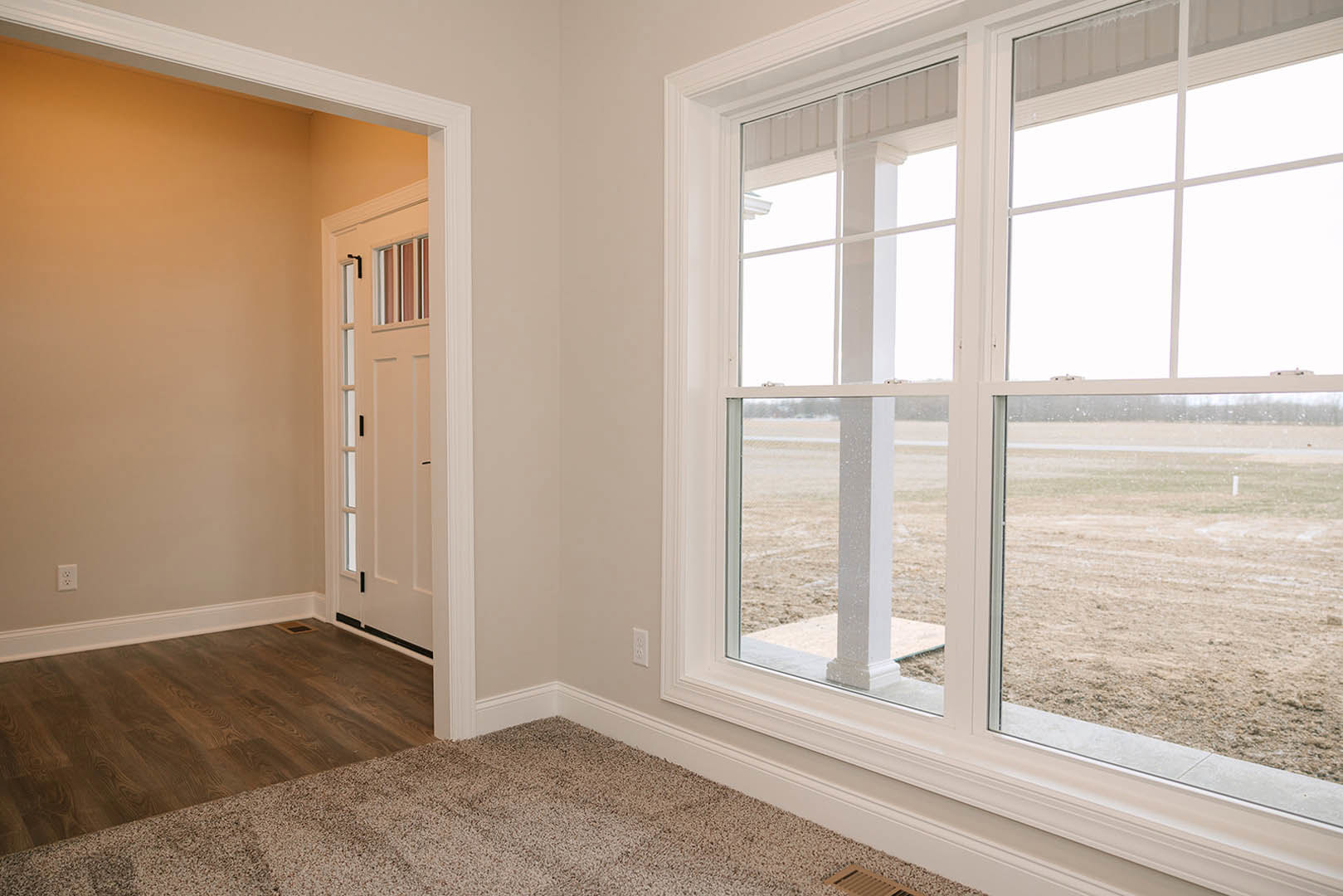 Spacious room featuring a large window overlooking a field, wood flooring, white walls with molding, and a door with carpeted threshold