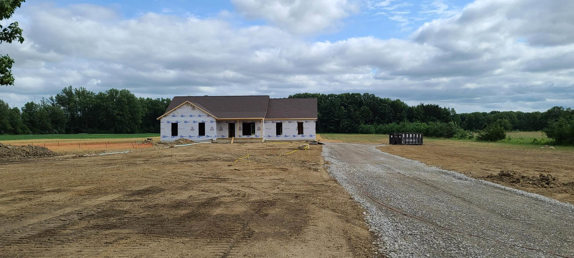 Partially built house with grey roof and exposed framing, surrounded by grassy field and trees, gravel driveway with black container, overcast sky