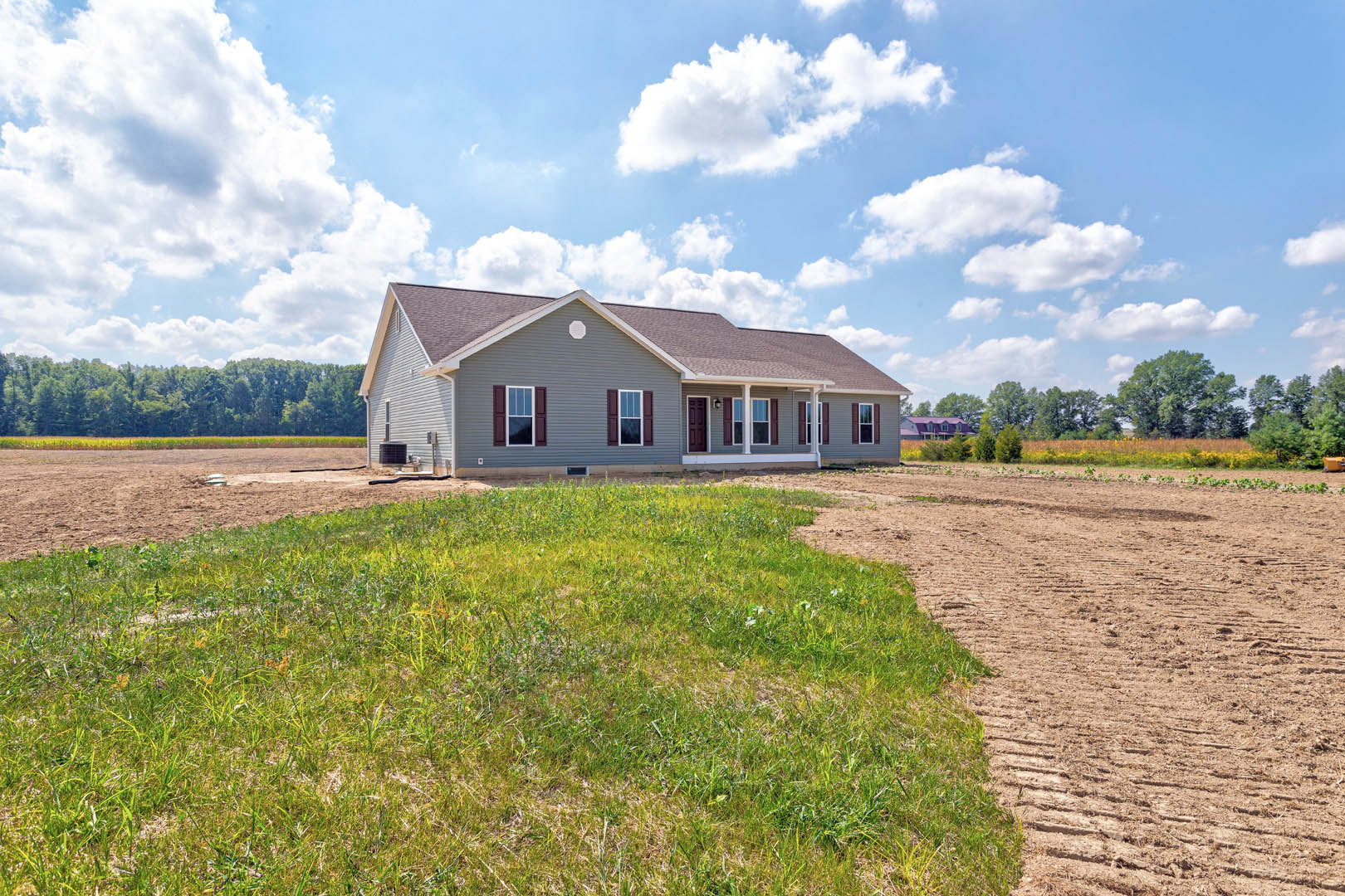 White-roofed house with light siding, dirt patch and tire tracks in front, grassy field, blue sky with clouds, trees in background