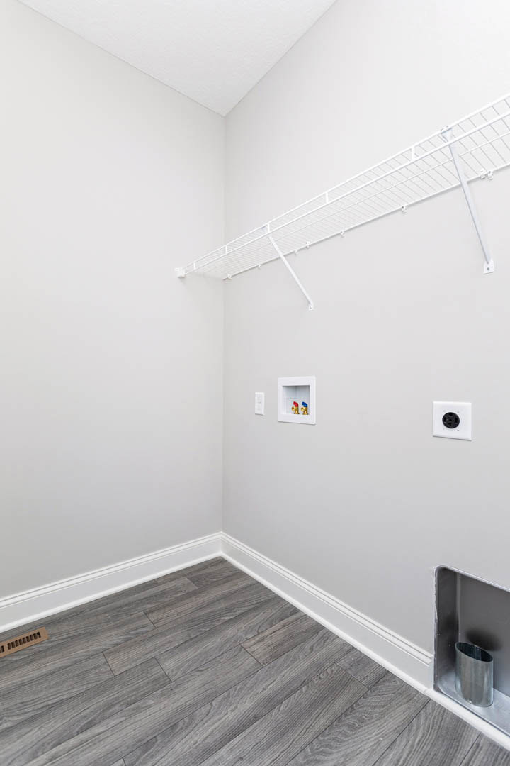 White floating shelf mounted on smooth white wall above wood floor with white baseboards, gold and blue faucets on white box, white electrical outlet with black circle, glass