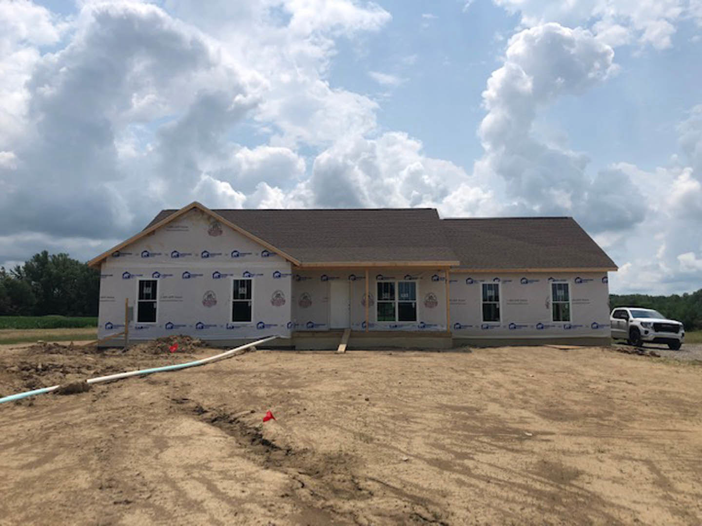 Framed house under construction with shingled roof, large dirt patch in foreground, white truck parked on road, red flag marker and pipe in dirt, cloudy sky overhead