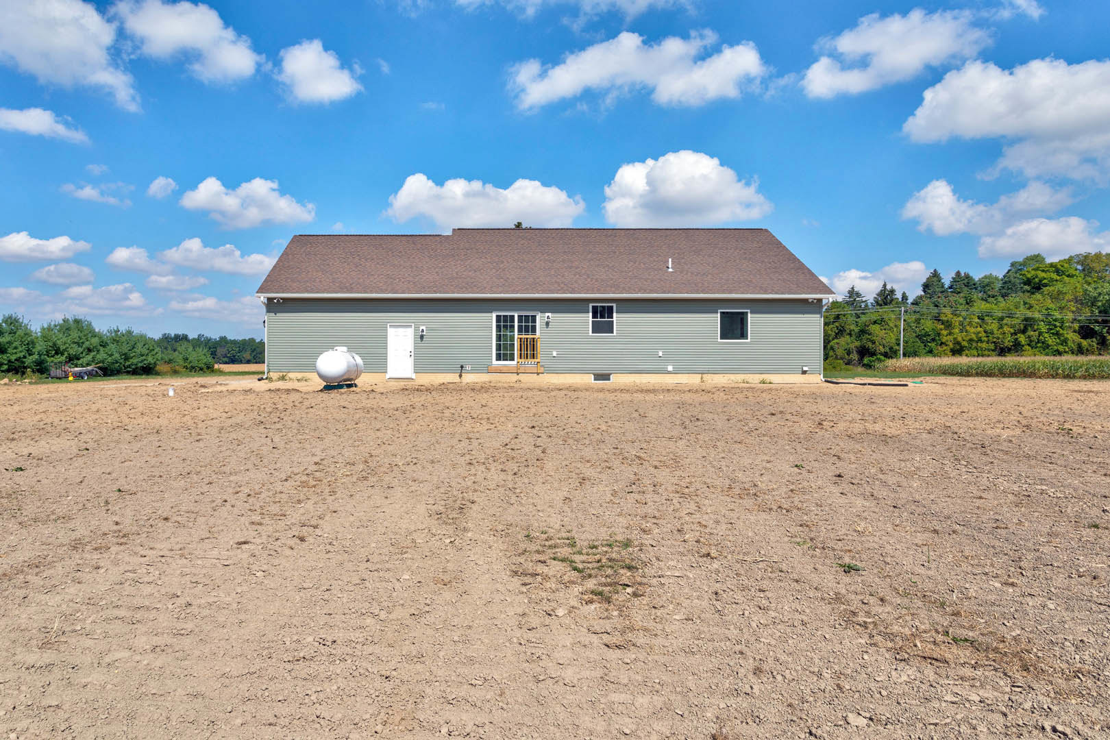 Single-story house with white-framed windows, covered porch, and white tank set on a dirt field under a cloudy sky
