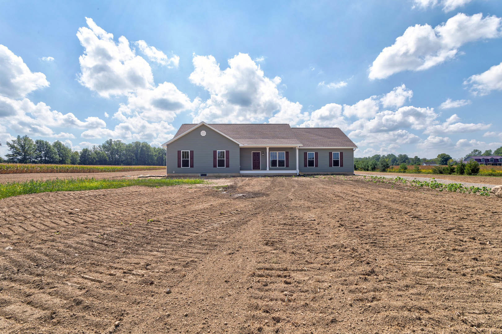 Grey house with white trim sits beside a dirt field marked with tire tracks, under a blue sky with scattered clouds; trees line the horizon.