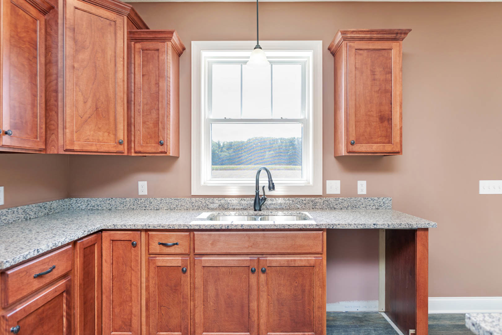 Granite countertop kitchen with stainless steel sink under a window, wooden cabinets and drawers, modern faucet, wall-mounted light fixture, and visible light switch.