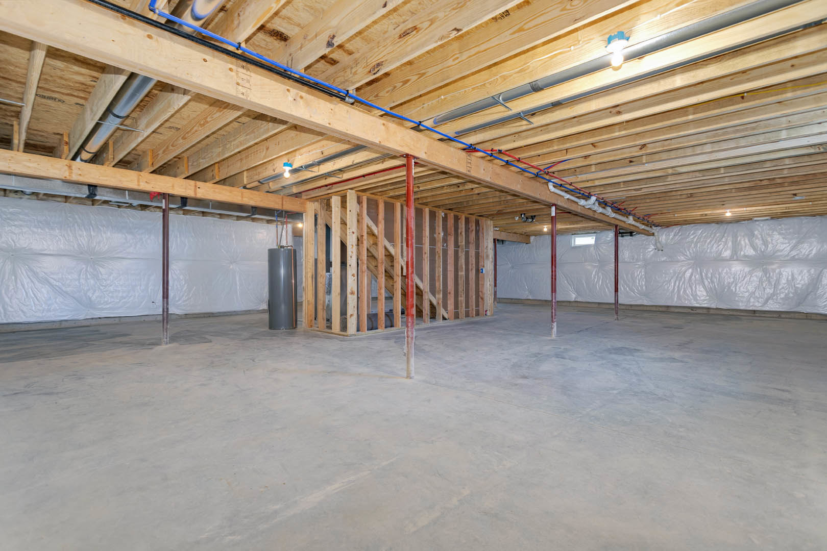 Basement room with exposed wood beams, blue and red pipes along the ceiling, concrete floor, grey cylinder, and white plastic sheeting on walls.