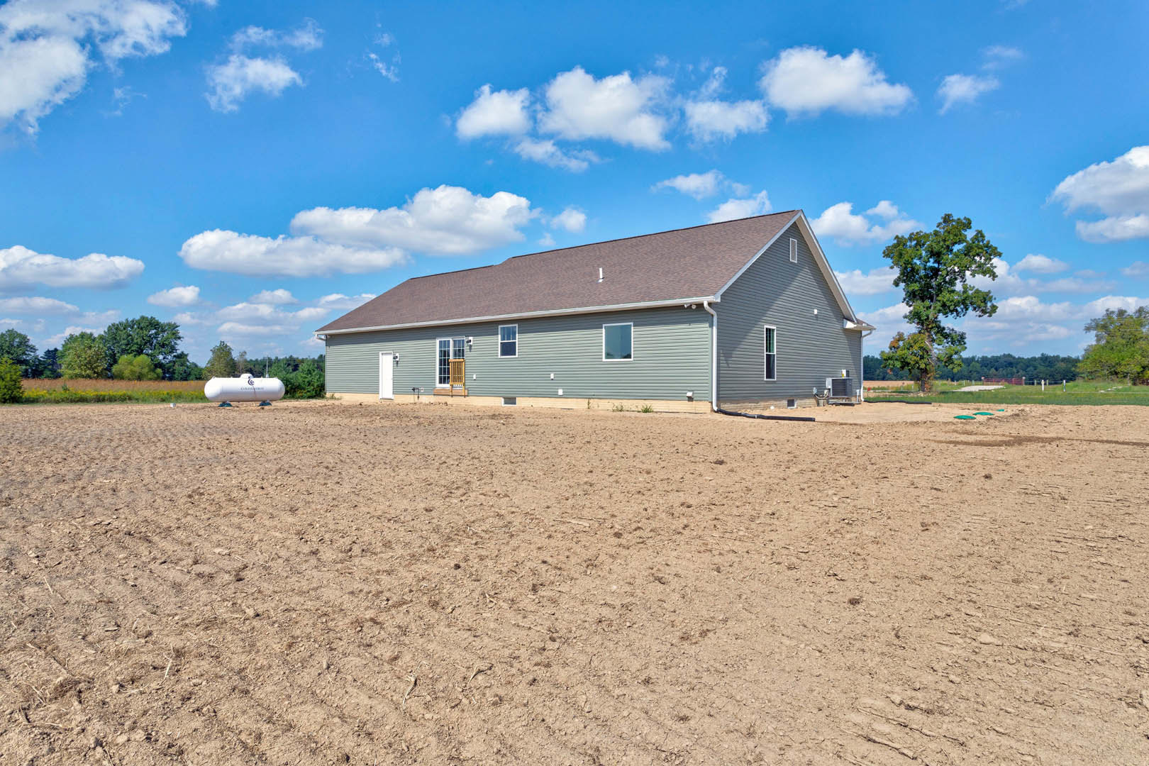 Single-story house with light siding and gabled roof, set beside a large dirt field, leafy tree in foreground, blue sky with scattered clouds overhead
