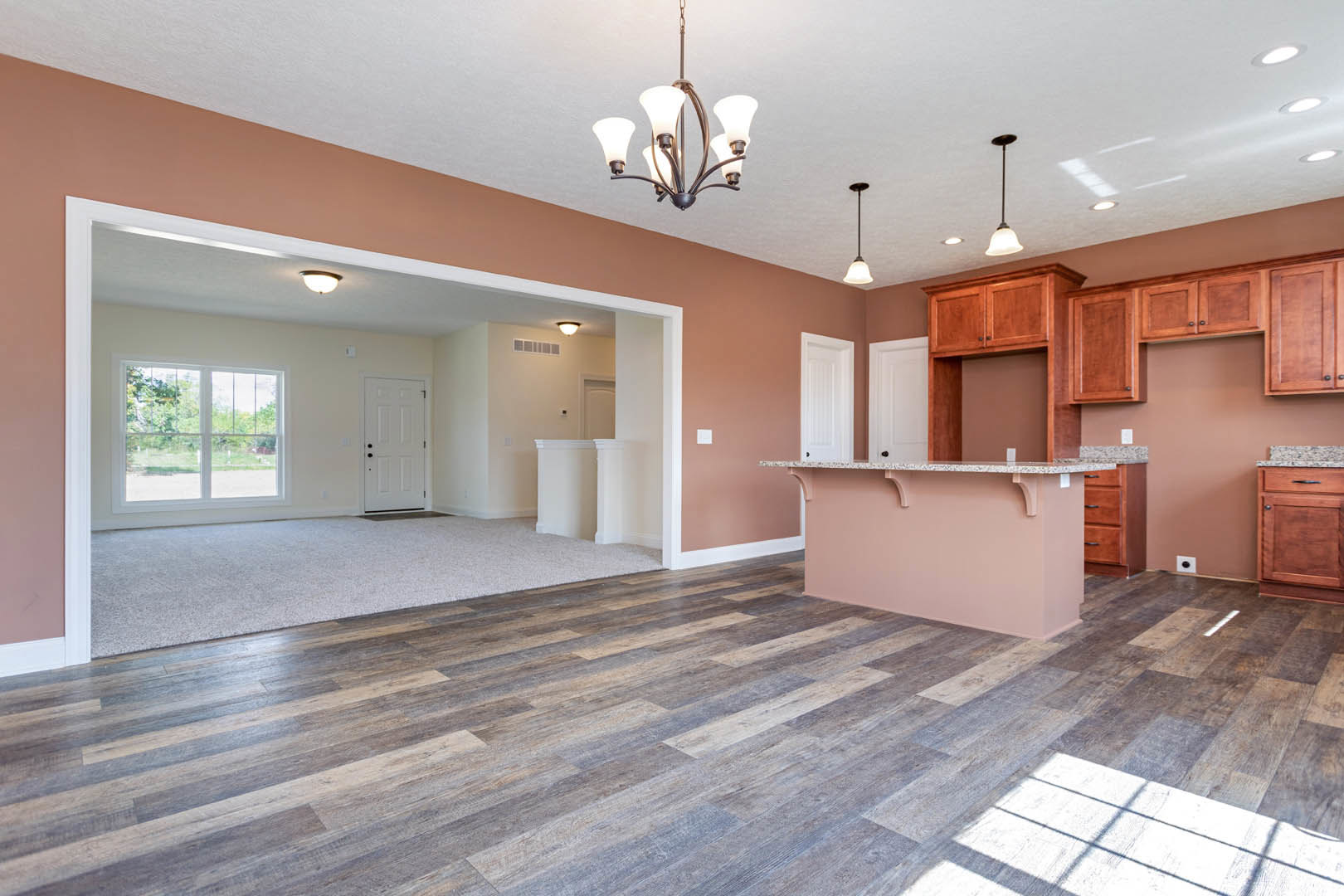 Open concept living room with wood flooring, pink accent wall, white kitchen cabinetry, black hardware, five-light chandelier, and window overlooking outdoor scenery