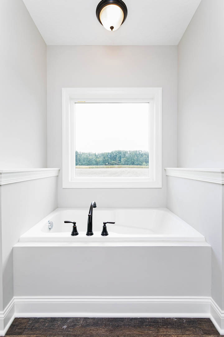Freestanding white bathtub beneath large window with tree views, chrome faucet, light fixture, and neutral-toned bathroom finishes