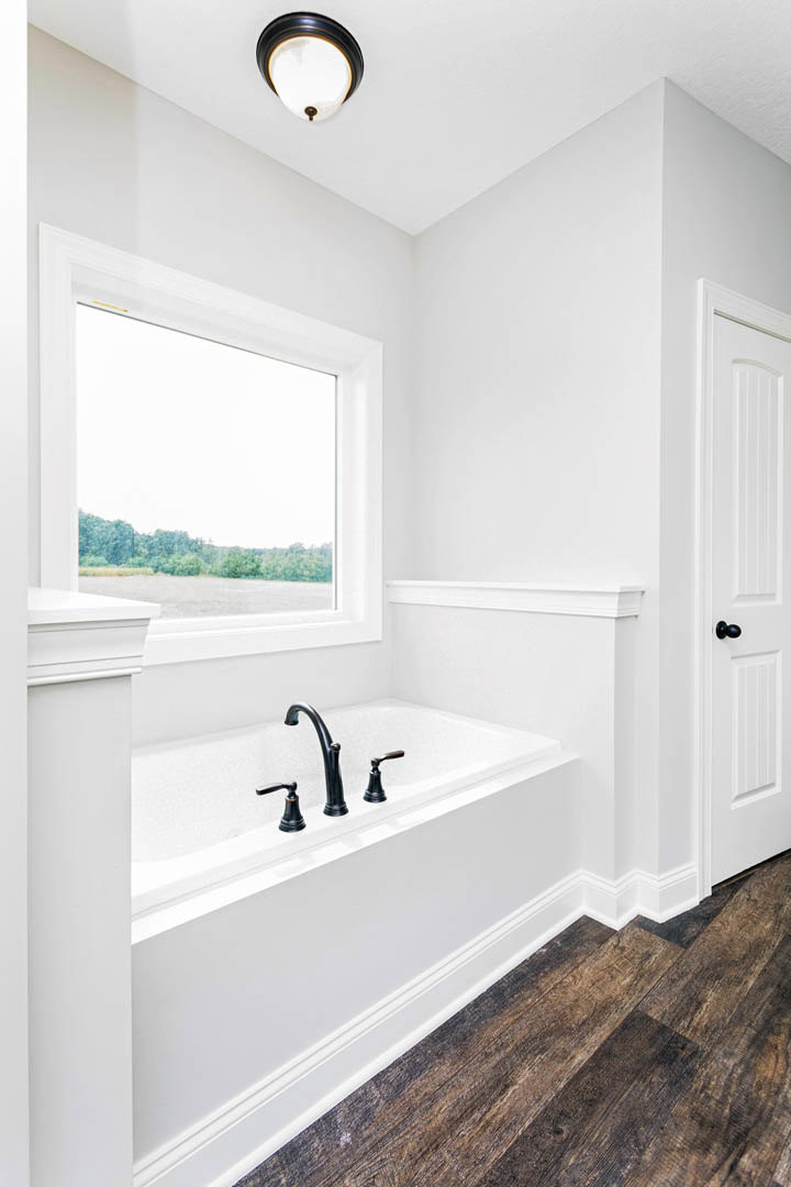 Freestanding bathtub beside large window with tree views, wood plank flooring, white walls, chrome faucet, and modern light fixture