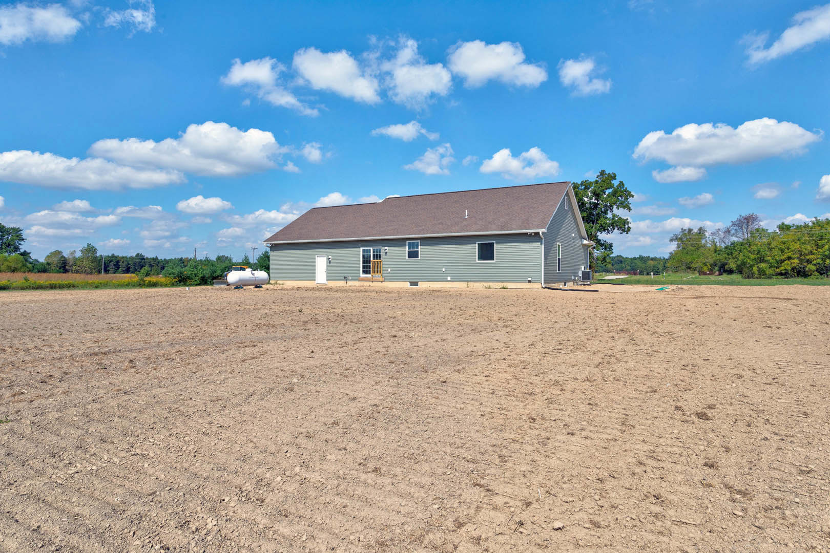 Single-story house with covered porch and white door, surrounded by a dirt field, trees in the background, and blue sky with scattered clouds