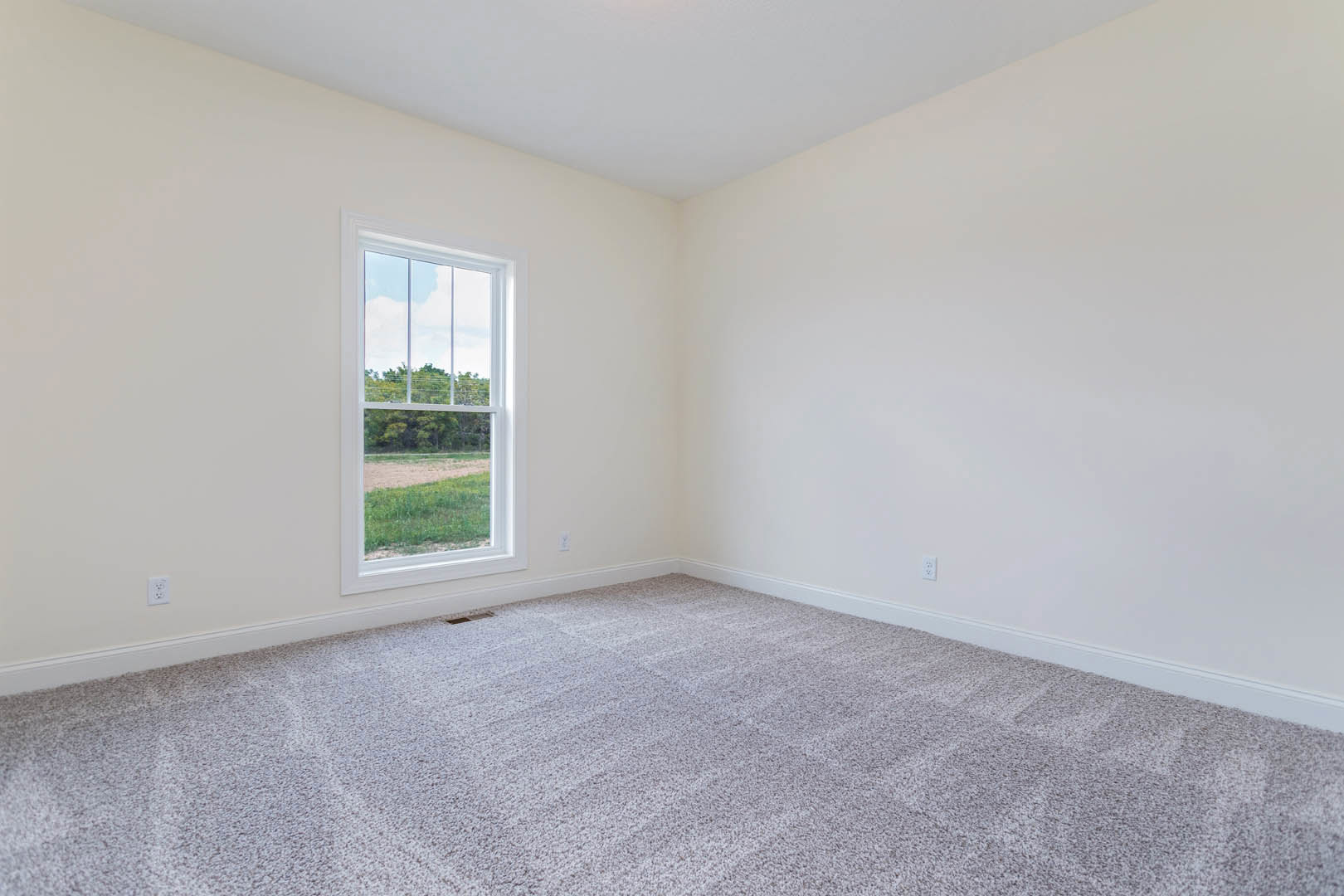 Bedroom with beige carpet, white walls and ceiling, large window overlooking grassy field and trees