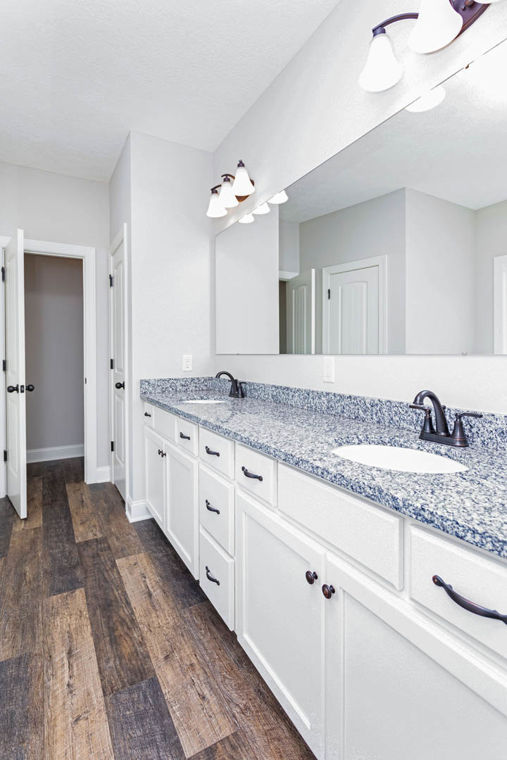 Bathroom with marble countertop, rectangular mirror above sink, chrome faucet, wood flooring, white cabinets, white door with black knobs, and modern light fixture.