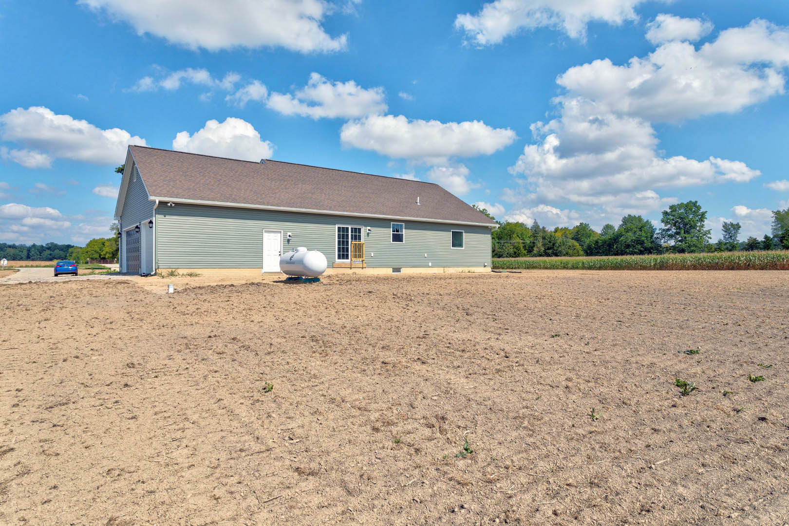 Single-story house with light siding, large white water tank in dirt field, green-leaved tree, blue sky with scattered clouds