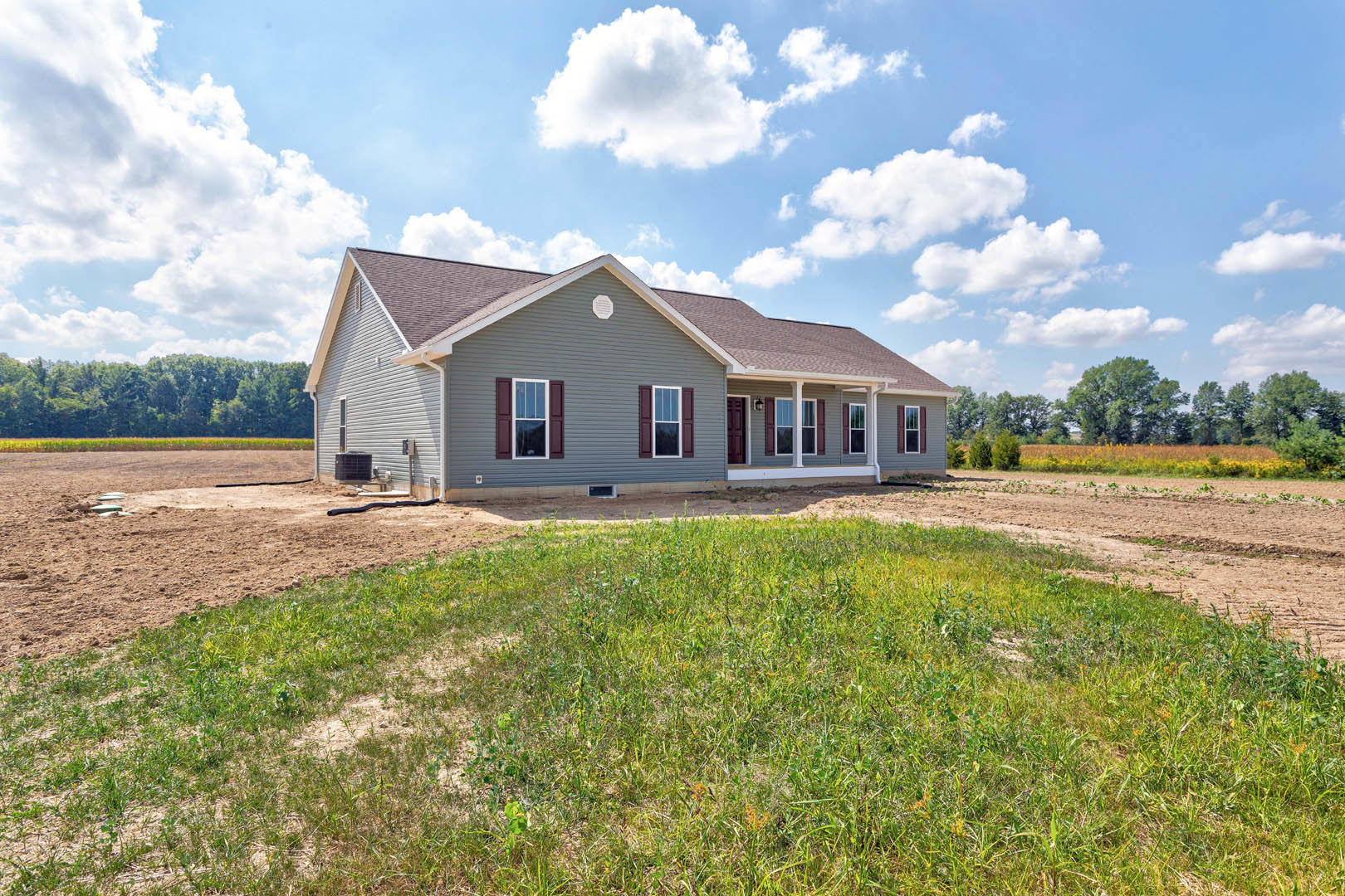 Two-story farmhouse with white siding, several windows, dirt field in foreground, grassy patches, and trees lining the background under a partly cloudy blue sky