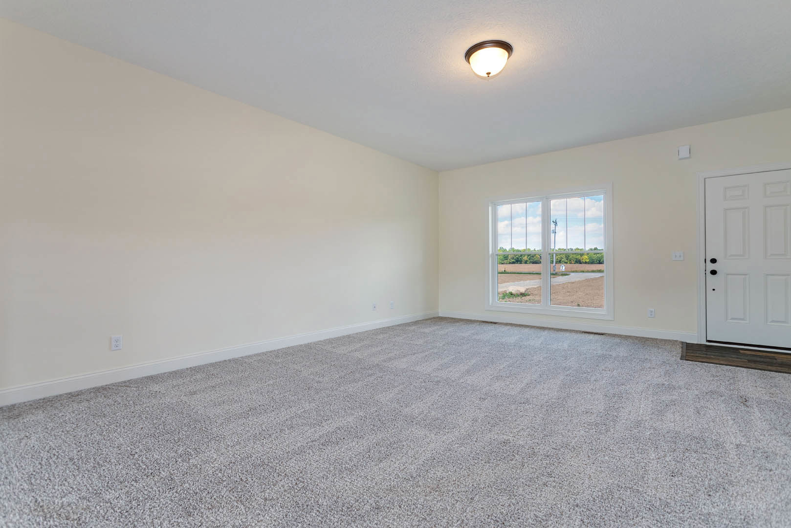 Neutral-toned carpeted room with white walls, white door featuring black knobs, ceiling-mounted light fixture, and large window overlooking grassy field under bright sky