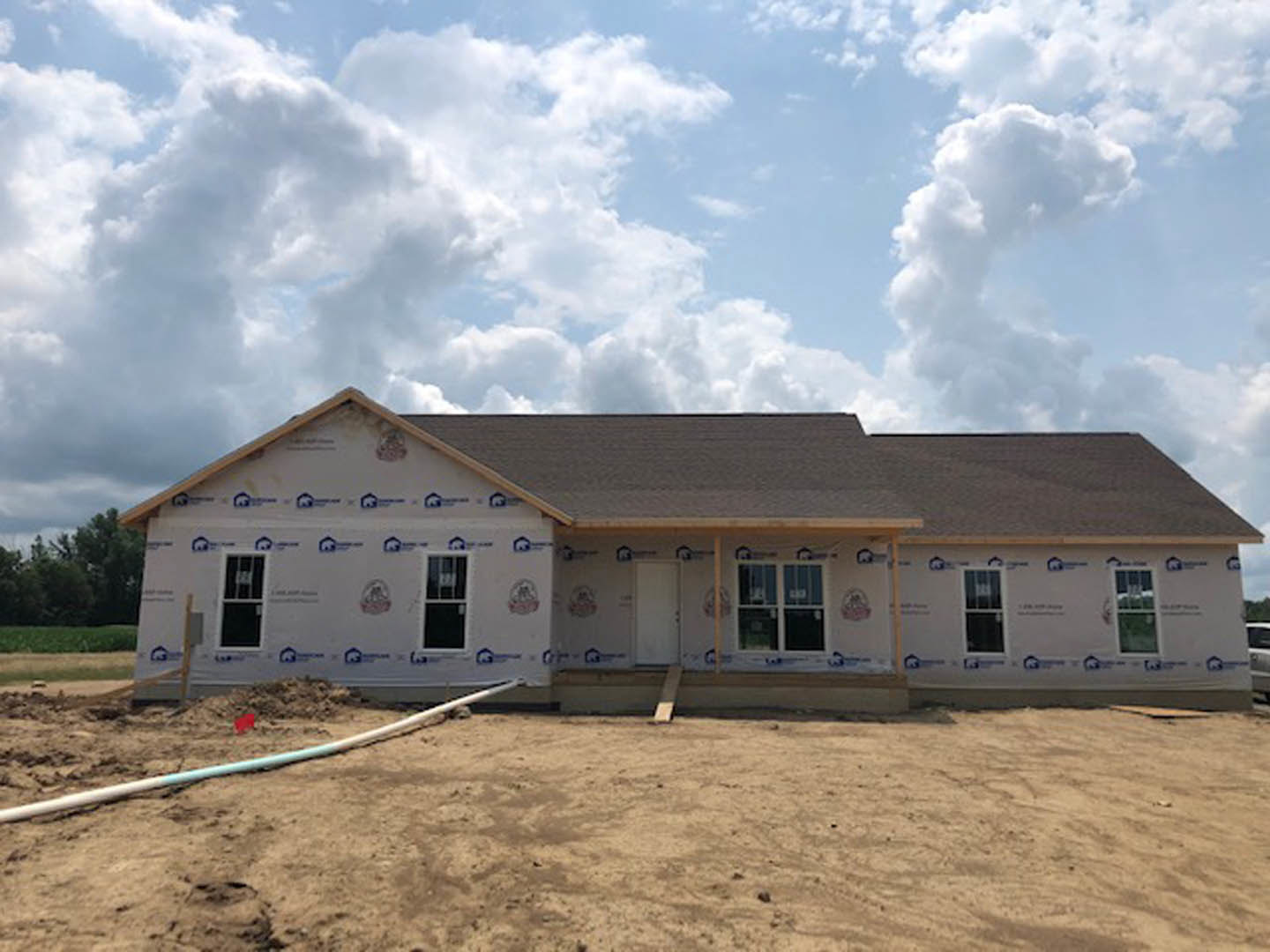 Framed house under construction with white walls, white door, exposed window, metal pipe on dirt ground, metal railing, and roof beneath blue sky with scattered clouds