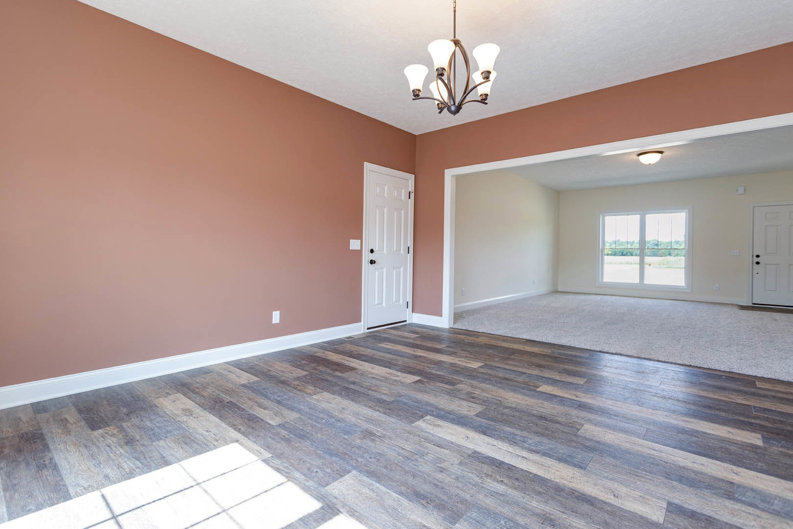 Wood floor with a white door featuring black knobs, five-light chandelier, window overlooking a field, carpeted area adjacent to hardwood flooring, white plaster walls and ceiling