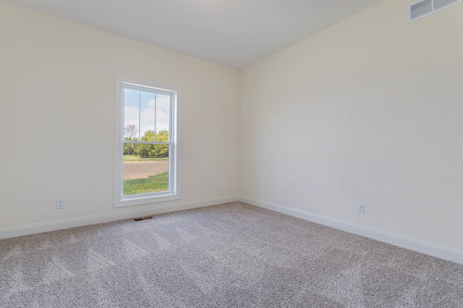 Carpeted room with large window overlooking trees and grassy area, white walls, natural light, outdoor view includes dirt road and cloudy sky.