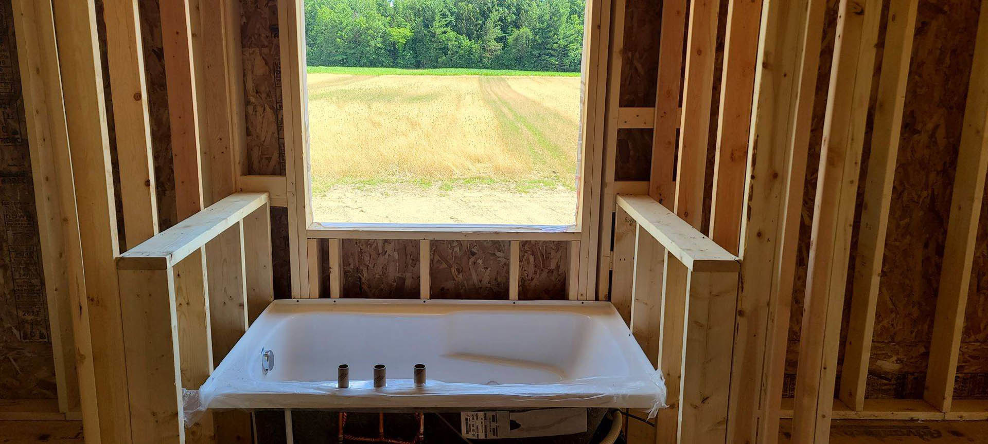Freestanding white bathtub beside large window overlooking grassy field and trees, wood-paneled walls, natural light filling indoor space